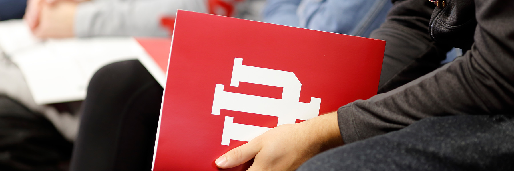 A seated person holds a red folder with a white IU Trident on it.